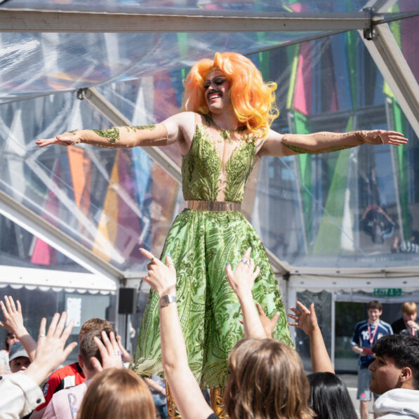 Dragqueen Mizz Privileze danser på et bord iført en grøn kjole og orange hår. Studerende sidder rundt om bordet og rækker deres hænder i vejret.