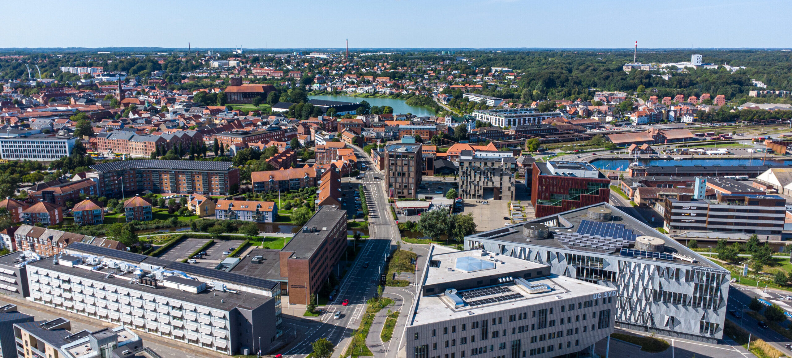 Koldingby og de fire uddannelsesinstitutioner ses oppefra på en solskinsfuld dag. Designskolen Kolding er den aflange bygning til venstre, efterfulgt at UC SYD og SDU som de to trekantet bygninger, og IBA er den brune bygning længere oppe. De er alle i gåafstand fra hinanden.