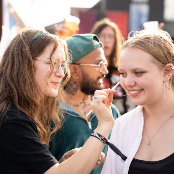 En ung kvinde får lagt farverig glitter makeup i ansigtet af en Campus Kolding studentermedhjælper.