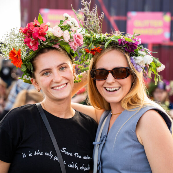 To unge kvinder smiler imod kameraet, imens de er iført farverige blomsterkranse.