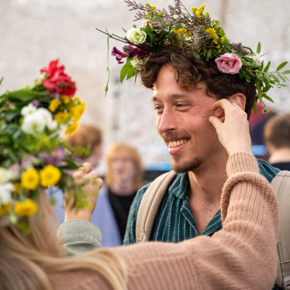 En ung mand er iført en farverig blomsterkrans og får glitter makeup i ansigtet.