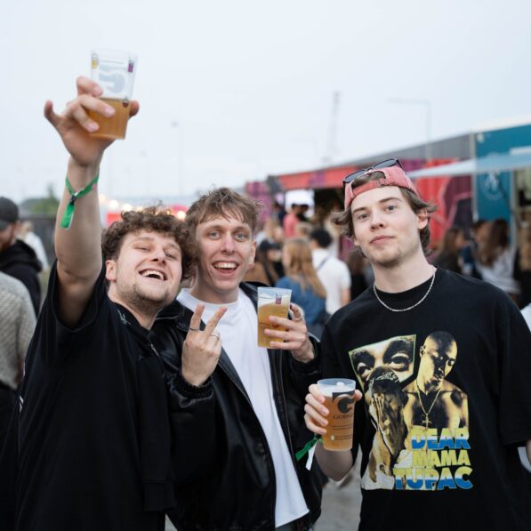 Tre unge mænd står sammen og smiler begejstret mod kameraet med oprakte øl.