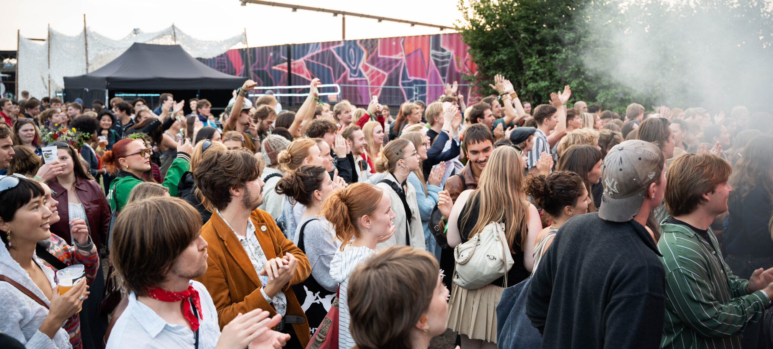 Et stort publikum af unge mennesker iført farverigt tøj står på festivalspladsen til Kontur 25 og kigger på mod scenen.