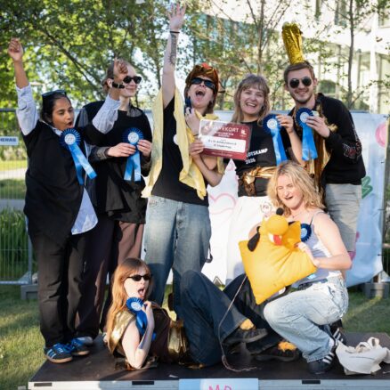 Vinderne af bedste kostume står samlet på et podie og viser begejstret deres præmier frem. Deres kostume er diverse stykker guldfarvet tøj.