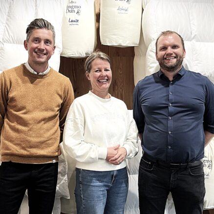 Tre personer står foran en væg fyldt med hvide dyner og puder, der er udstillet i et showroom.