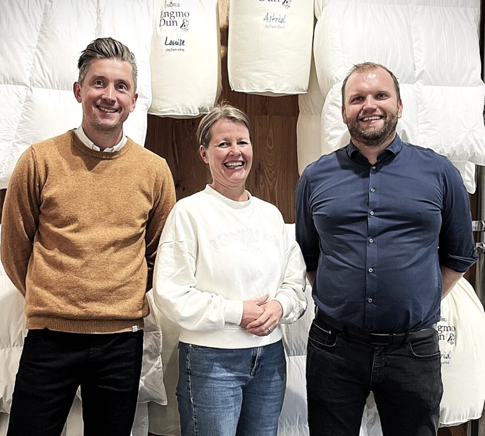 Tre personer står foran en væg fyldt med hvide dyner og puder, der er udstillet i et showroom.