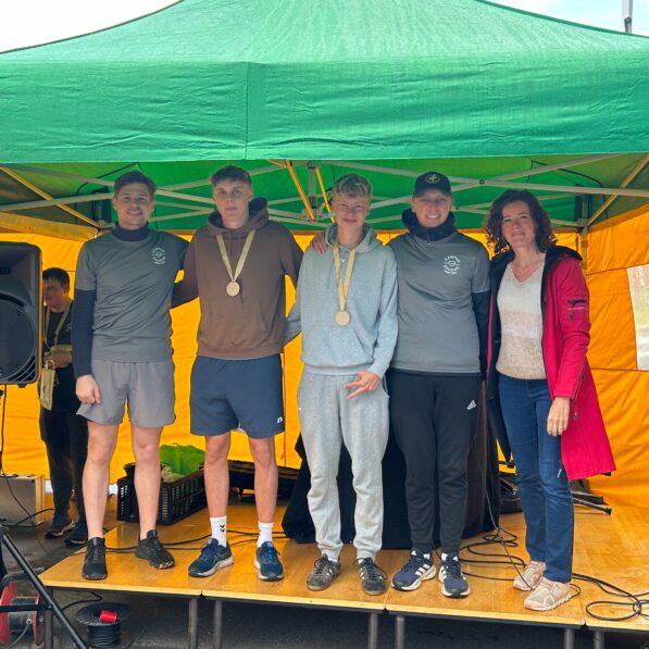 To unge mænd iført Campus Kolding Løbets træ-medaljer står på et podium og smiler mod kameraet sammen med 3 andre.