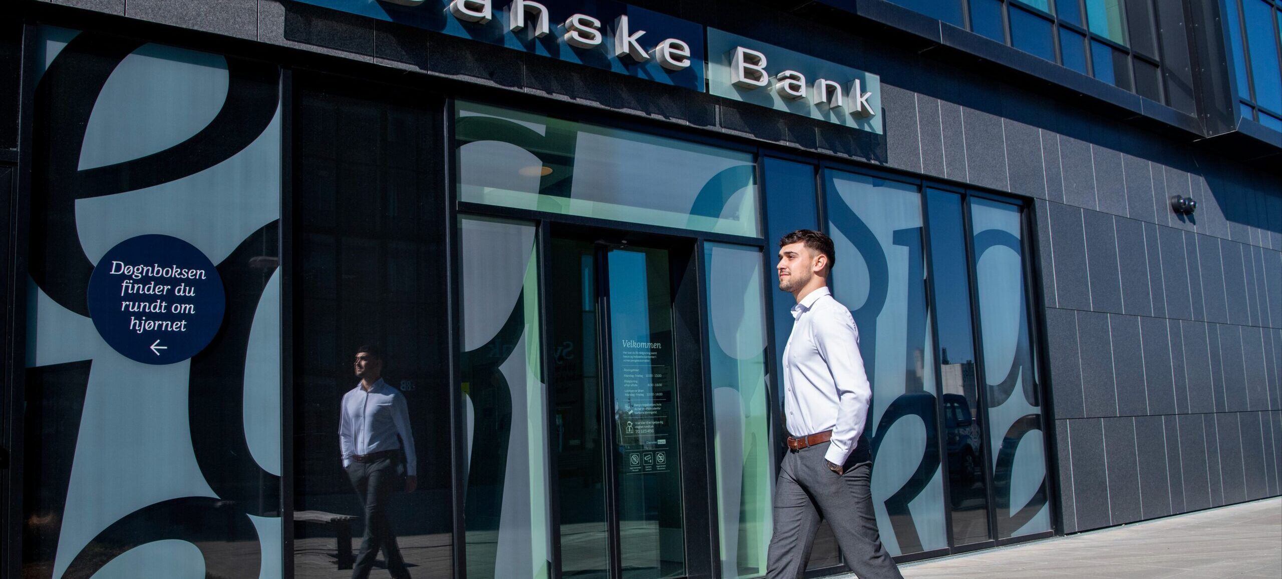 Mergim er iført skjorte og bukser, og han er på vej ind i en bygning med skiltet "Danske Bank" over indgangen.