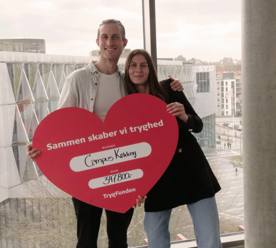 Klaus og Amalie-Lærke står smilende med armene rundt om hinanden og et stort rødt pap-hjerte med donationen fra TrygFonden. Hjertet har titlen "Sammen skaber vi Tryghed" adresseret til Campus Kolding med 347.800 kr.