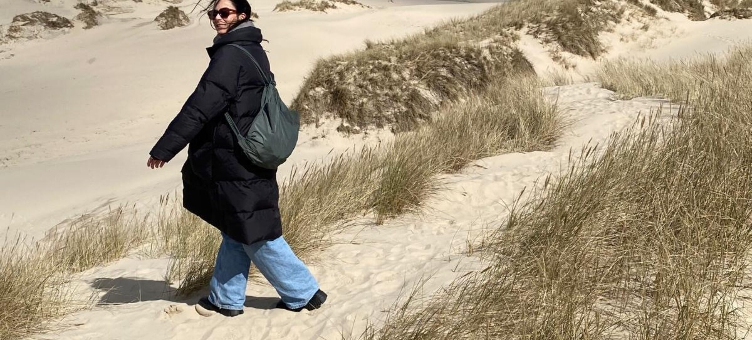 Sara Bilić går på stranden med vind i håret og varm jakke. Hun kigger tilbage mod kameraet og smiler.