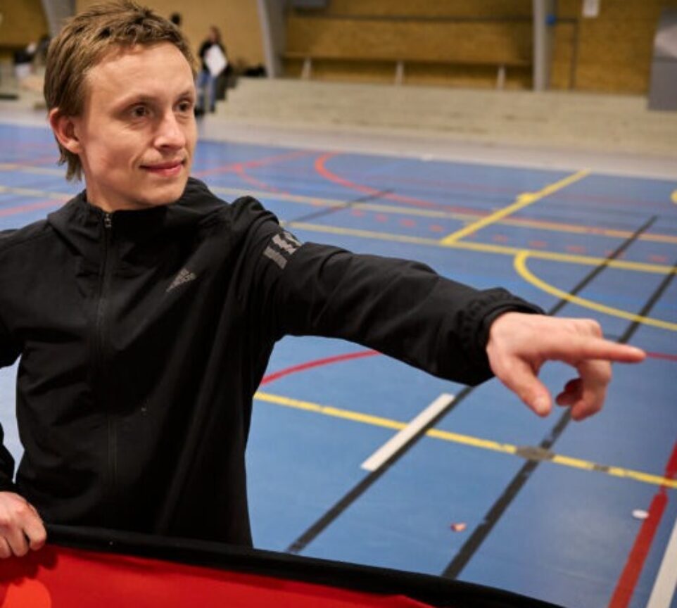 Oliver står i en indendørs fodboldhal med noget rødt i hånden, imens han peger på noget udenfor billedet.