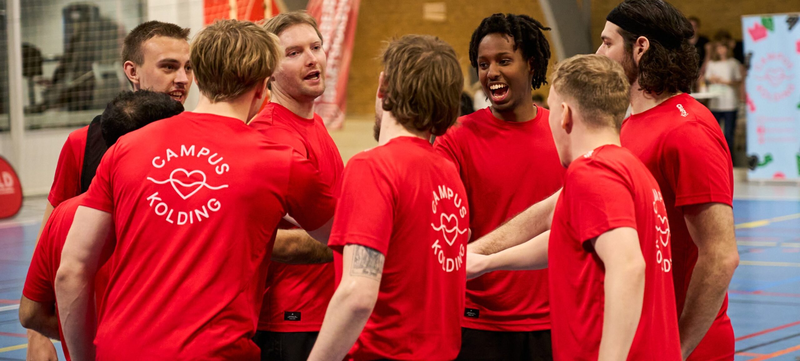 En flok unge mænd står i en indendørs sportshal med deres hænder samlet. De er iført røde Campus Kolding T-shirts.