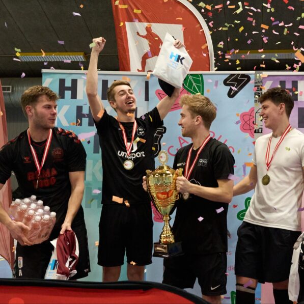 Fem unge mænd står jubelende med medaljer om halsen og en pokal i hænderne, mens konfetti festligt daler ned fra loftet.