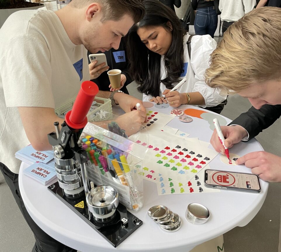 3 studerende står bøjet over en stand og laver badges med farverigt papir, klistermærker og tusser.