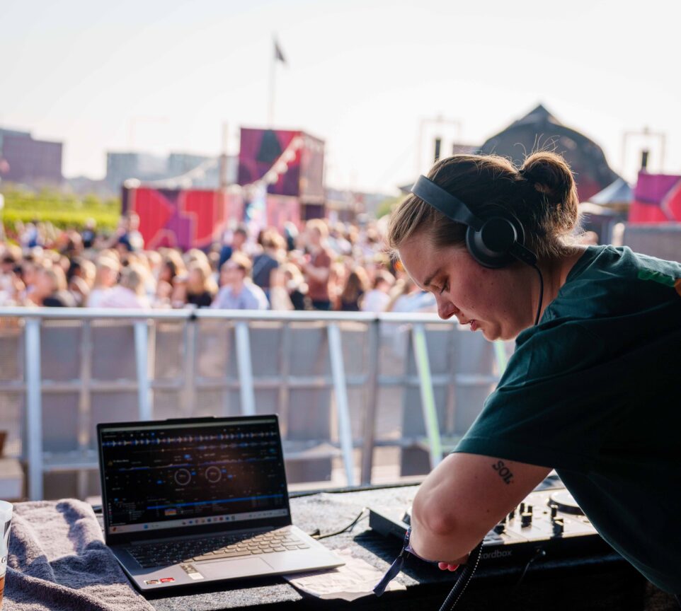 Linnea står koncentreret over en DJ stand med høretelefoner og computer. Bagved hende står publikummet til KONTUR festivalen.