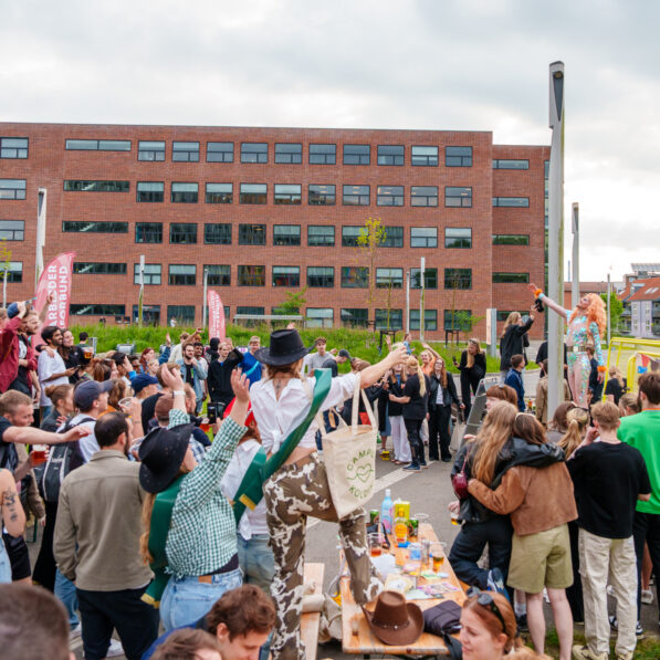 Campus pladsen er fyldt med studerende, der alle er iført farverige kostumer og begejstret rækker hænderne i vejret og synger. På et bord står Mizz Privileze og synger for.